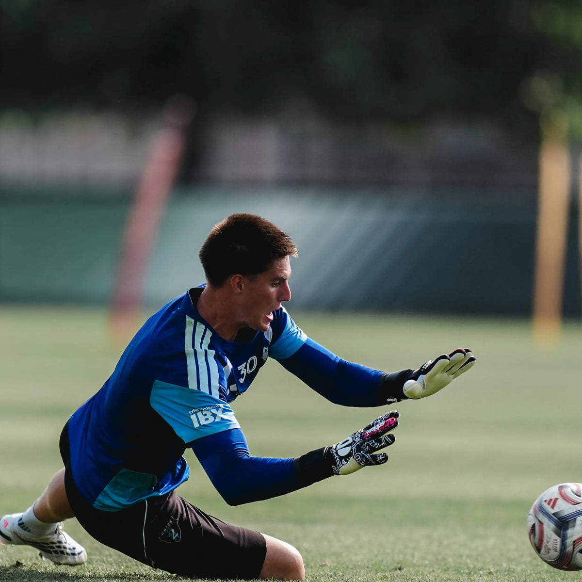 A soccer goalkeeper in a blue and black long-sleeve jersey with white gloves dives on a green grass field toward a soccer ball.